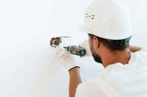 A worker in a white hard hat and gloves using a drill on a wall outlet.